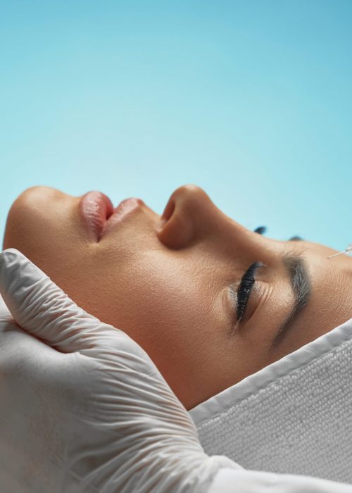 Close up of botox injection in female forehead. Side view of cosmetologist using syringe with special liquid, holding chin while patient with towel on head lying. Concept of cosmetology, beauty.